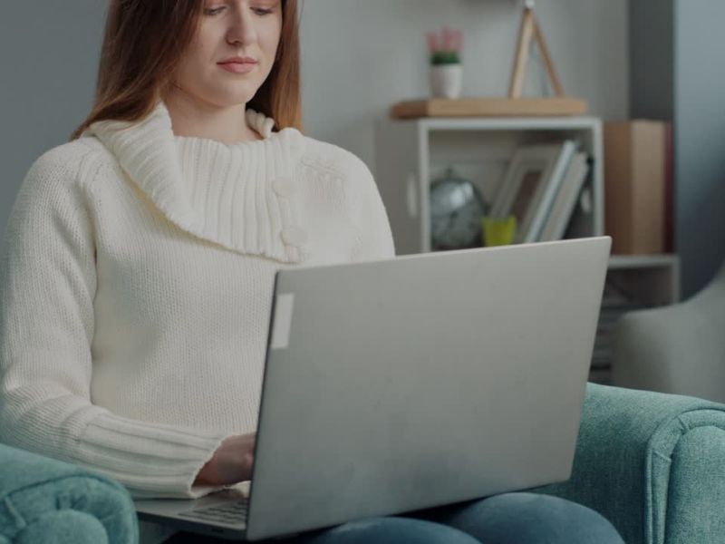 Devojka smeđe kose u belom džemperu sedi u plavoj fotelji i koristi laptop