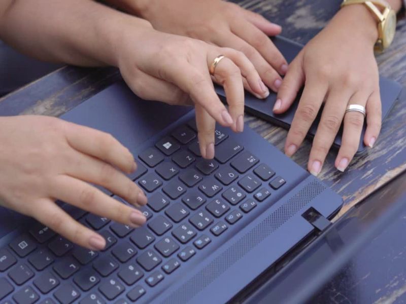 Dve osobe seda za laptopom i pretražuju nešto, laptop stoji na drvenoj površini a telefon je na stolu