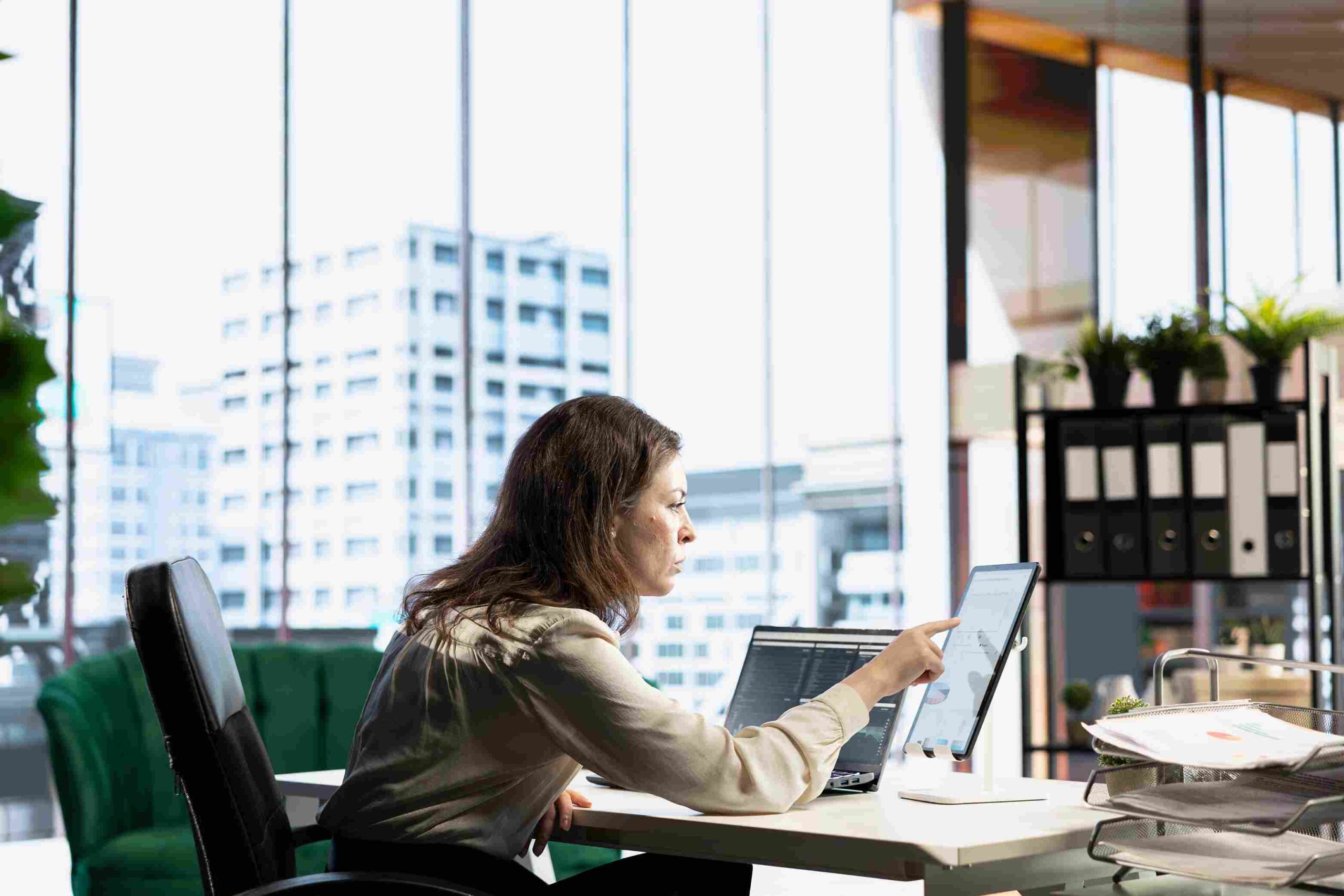 Žena u kancelariji koristi tablet i laptop za analizu podataka