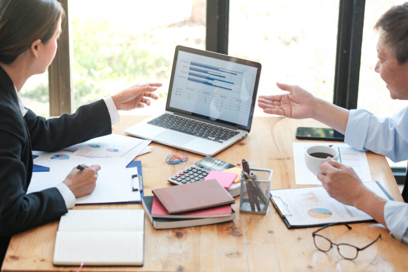 Dvoje kolega analiziraju finansijske izveštaje na laptopu uz grafikone i dokumente na stolu