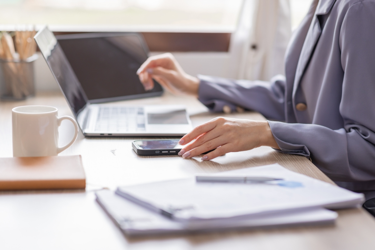 osoba koristi telefon, pored nje laptop i dokumenti