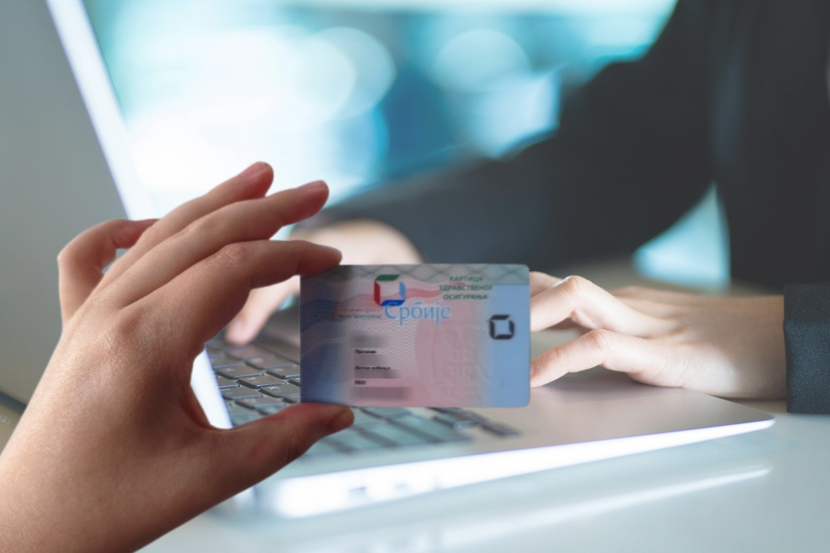 osoba koristi laptop i druga osoba drži zdravstvenu knjižicu u ruci