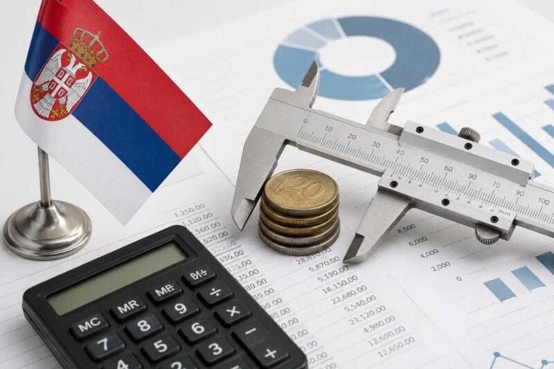 Šubler, novčići i kalkulator na finansijskim dokumentima uz zastavu Srbije.